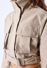 Beige cropped jas gemaakt van gestructureerde stof met twee voorzakken, hoge kraag en een verstelbare gesp aan de onderkant. Heeft subtiele stikdetails.