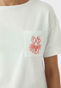 T-shirt en coton blanc avec une poche poitrine brodée d'un homard orange. Manches courtes et encolure ronde. Texture lisse.