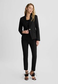 Zwarte getailleerde blazer met een enkele gouden knoop, gecombineerd met slim-fit zwarte broeken en zwarte sandalen met verenaccenten.