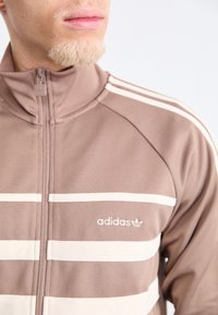 Chaqueta de chándal marrón con cremallera, que presenta rayas horizontales blancas, una textura suave y un logo de adidas en la parte frontal izquierda.