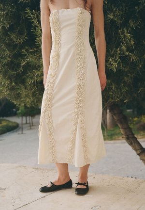 Donna che indossa un vestito crema senza spalline con dettagli di volant verticali e scarpe basse nere Mary Jane, in piedi all'aperto vicino agli alberi.