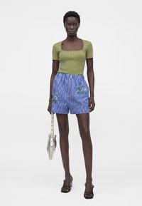 Femme portant un haut vert à encolure carrée et manches courtes, un short fleuri rayé bleu et blanc, des sandales noires à talons, tenant un sac à franges argenté.