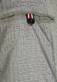 Tejido gris con un patrón de texto repetido. Presenta un cierre de botón y un acento a rayas en rojo, blanco y azul en la parte superior de la solapa.