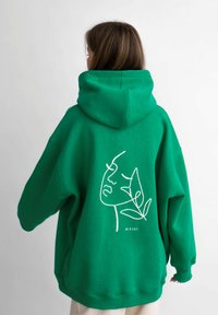 Felpa verde oversize realizzata in tessuto morbido, con un design di viso in line art e foglia bianca sul retro. Cappuccio ampio e vestibilità comoda.