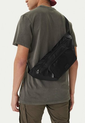 Man wearing olive green t-shirt and pants with black crossbody sling bag and red cap, shown from the back against white background.