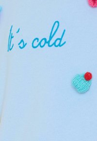 Jasnoniebieski sweter z haftowanym napisem "Zimno" w turkusowym kolorze oraz dzianinowym detalem w kształcie babeczki z czerwoną wisienką z boku.