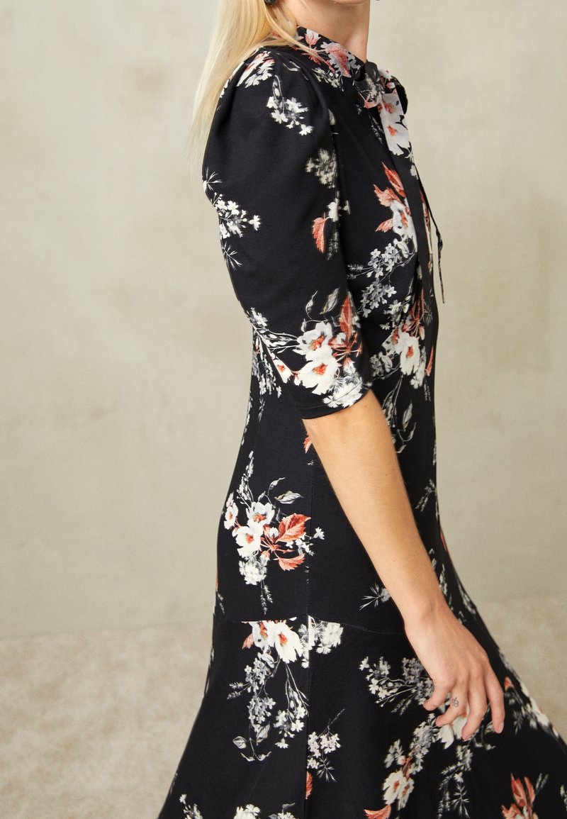 Femme portant une robe noire cintrée avec des motifs floraux blancs et orange, des manches bouffantes jusqu'au coude et un col noué.