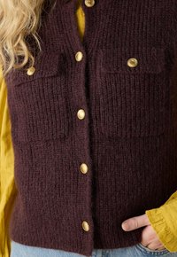 Bordeaux gebreide vest met geribbelde textuur, voorzien van twee borstzakken en gouden knopen. Draag het over een gele blouse met lange mouwen.