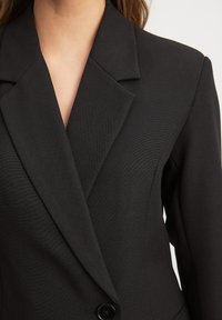 Melns žaketis ar izteiktu apkakli, vienu pogu, strukturētu piegriezumu un teksturētu audumu. Tuvplāns, akcentējot lakatu un apkakles detaļas.