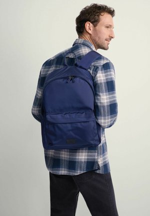 Granatowy plecak z gładkiego, trwałego materiału; posiada dwie komory na zamek oraz przednią kieszeń; noszony na koszuli w kratę.