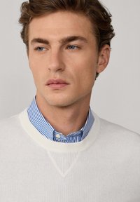 Witte ribgebreide trui voor mannen met een V-hals, gedragen over een blauw-wit gestreept overhemdd. Close-up van de details van de halslijn.
