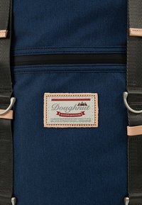 Sac à dos en tissu bleu avec un patch en cuir beige affichant le nom de la marque. Il présente des bretelles grises, une fermeture éclair noire et des accents métalliques argentés.