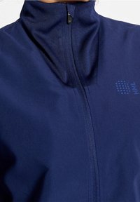 Marinblå zip-up jacka med hög krage, slät textur och subtil logotypdetalj nära dragkedjan. Tillverkad av ett lätt material.
