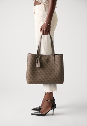 Brown patterned tote bag with dual handles, a zipper closure, and a decorative tag. Paired with cream pants and shiny black heels.