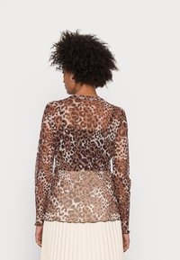 Blusa leve e transparente de mangas longas com padrão de leopardos castanho e bege. Apresenta um decote arredondado e uma bainha com babados. Tecido texturizado com uma aparência leve.