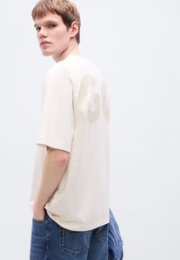 Κοντομάνικη μπλούζα σε κρεμ απόχρωση με μεγάλο, ανοιχτόχρωμο τύπωμα "GO" στην πλάτη. Το ύφασμα φαίνεται μαλακό με άνετη εφαρμογή.