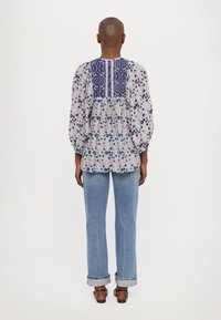 Blumenbluse in Weiß mit blauen und roten Akzenten, ausgestattet mit einem marineblauen gemusterten Einsätze, Puffärmeln und einem lässigen Schnitt, kombiniert mit hellblauen Jeans.