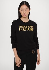 Melna džemperis ar zelta "VERSACE JEANS COUTURE" tekstu. Izceļas ar apaļu kaklu, ribotām piedurknēm un klasisku, pieguļošu dizainu.