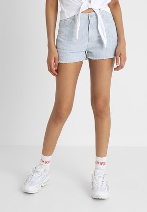 Frau trägt hellblaue, gestreifte Jeansshorts, weißes, vorne geknotetes Hemd, weiße Socken mit rotem Text und weiße Turnschuhe, steht mit gespreizten Beinen.