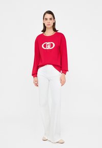 Felpa rossa con logo bianco ricamato, abbinata a pantaloni bianchi a palazzo e scarpe chiare. Tessuto morbido e vestibilità rilassata.