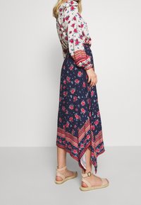 Robe fleurie à manches longues, base blanche, fleurs rouges, jupe marine, ourlet asymétrique, accents à motifs. Associée à des sandales espadrilles beige.