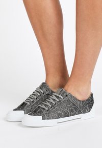 Grijze canvas sneakers met patroon, voorzien van witte rubberen zolen en veters. Het bovenwerk heeft een gestructureerd ontwerp en een subtiel merkdetail.