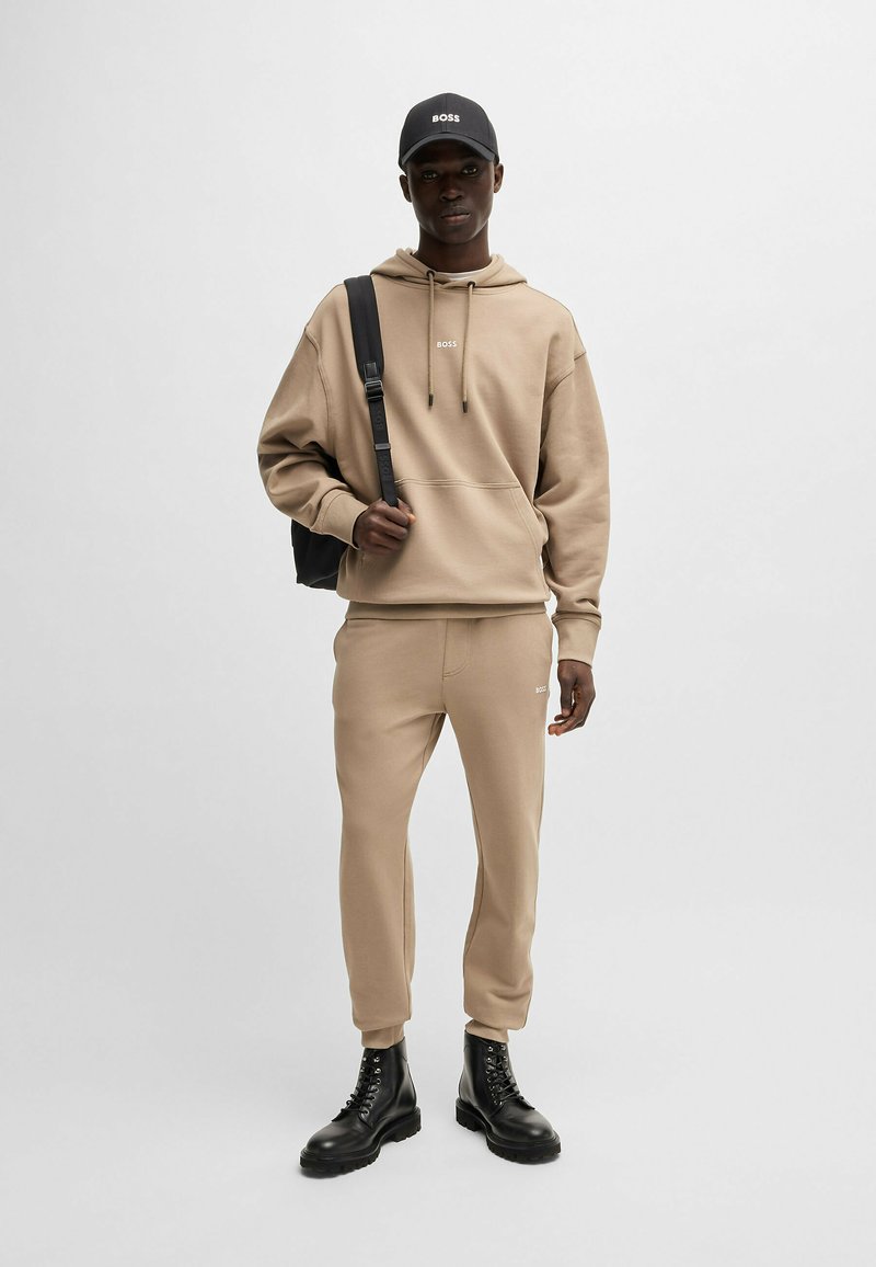 Beige hoodie with adjustable drawstring, logo detail. Matching joggers, tapered fit. Black backpack and chunky black boots. Smooth texture.