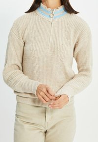 Beige Strickpullover mit einem Viertelreißverschlusskragen, der eine hellblaue gerippte Akzentuierung am Kragen und ein strukturiertes Material aufweist. Lange Ärmel, elastische Bündchen.