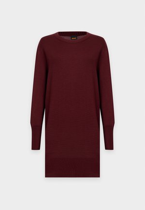 Femme aux longs cheveux noirs portant une robe-pull ample à manches longues de couleur bordeaux foncé et des chaussures à talons noirs, debout devant un fond uni.