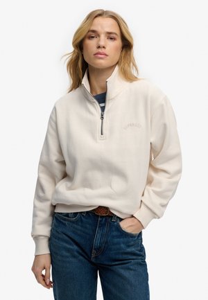 Donna con capelli biondi che indossa una felpa crema con cerniera a quarto giro sopra una maglia a righe, jeans blu con cintura intrecciata marrone, mano in tasca.