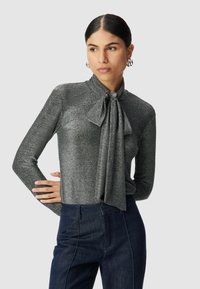 Blusa grigia a maniche lunghe in tessuto lucido con fiocco legato al collo, abbinata a pantaloni jeans scuri a vita alta.