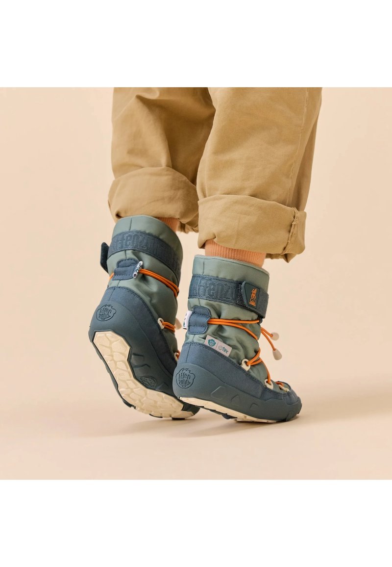 Enfant portant des bottes d'hiver isolées bleues et vertes avec des lacets orange et un pantalon beige avec les revers retroussés sur un fond neutre.