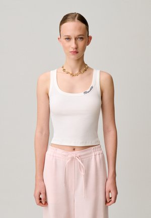 Joven con el cabello peinado hacia atrás, lleva un crop top blanco sin mangas con el texto "Claudie" y pantalones con cordón de color rosa claro.