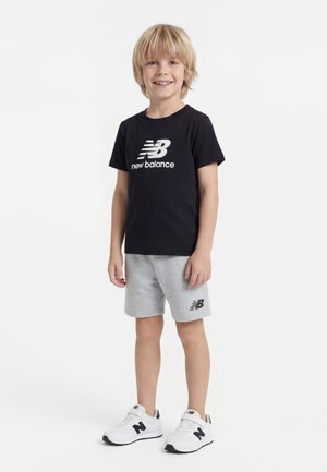 Ragazzo giovane in piedi, indossa una maglietta nera New Balance, pantaloncini grigi con logo e scarpe da ginnastica bianche New Balance, sorride alla fotocamera.