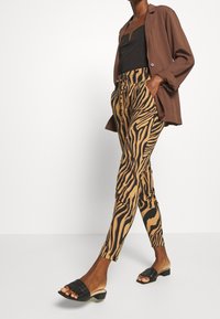 Pantalons à rayures zèbre marron et noir, en tissu léger. Associés à un haut noir et un blazer marron. Sandales tressées noires.