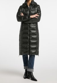 Osoba v dlouhé černé péřové bundě se zipem vpředu, s ohrnutými modrými džíny a černými kotníkovými botami na podpatku, se založenýma rukama na bílem pozadí.