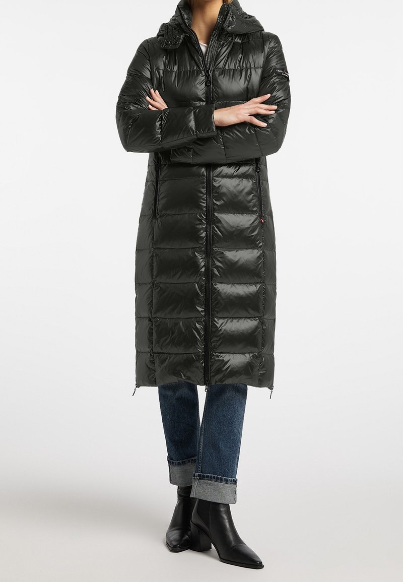 Osoba v dlouhé černé péřové bundě se zipem vpředu, s ohrnutými modrými džíny a černými kotníkovými botami na podpatku, se založenýma rukama na bílem pozadí.