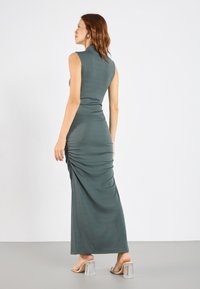 Maxiklänning i mörkgrön ribbad textil med hög hals och rynkade detaljer på sidan; kombinerad med sandaler med klack i genomskinligt material.