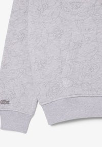 Felpa grigia con motivo a rilievo floreale, polsini e orlo a coste. Presenta il logo Lacoste ricamato in filo scuro sul polsino inferiore.