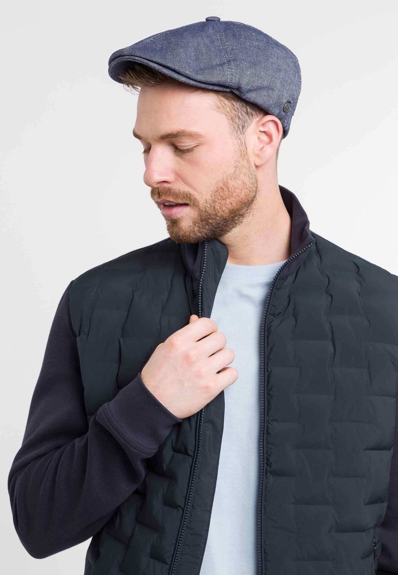 Men's dark vest with quilted texture, worn over a light blue shirt. A flat cap in denim fabric completes the outfit. Casual design with zipper details.