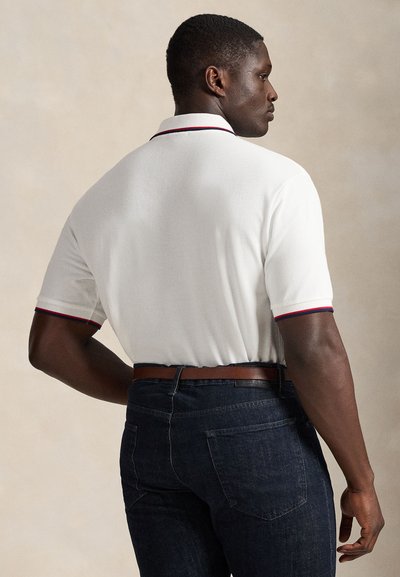 Bărbat în polo alb cu mâneci scurte cu bordură roșie și bleumarin, blugi închiși la culoare și curea maro, văzut din spate pe un fundal neutru.