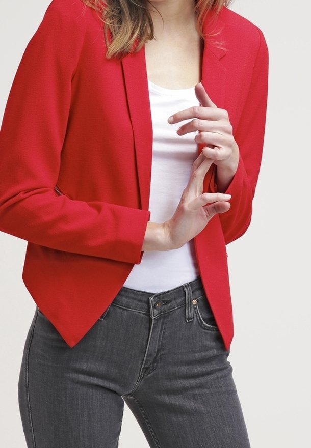 Personne portant un blazer rouge et une chemise blanche, qui croise les doigts près de la taille, portant un jean gris foncé visible des hanches vers le bas.