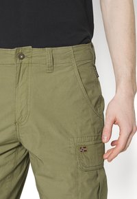 Pantaloni cargo verde olivo in tessuto testurizzato, con diverse tasche e una toppa decorativa che presenta una piccola bandiera sul lato.