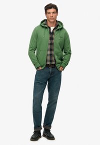 Hombre con sudadera verde con cremallera sobre una camisa de cuadros, vaqueros azules con dobladillos, botas negras, de pie con las manos en los bolsillos sobre un fondo blanco.