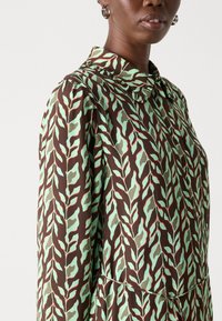 Camicia a maniche lunghe con colletto, realizzata in un tessuto marrone decorato con un motivo di foglie verdi e accenti rossi, con una texture liscia.