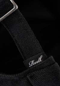 Correa de tela negra con una superficie texturizada, que presenta una hebilla de metal plateado y una etiqueta cosida que dice "Reell" en una fuente cursiva blanca.