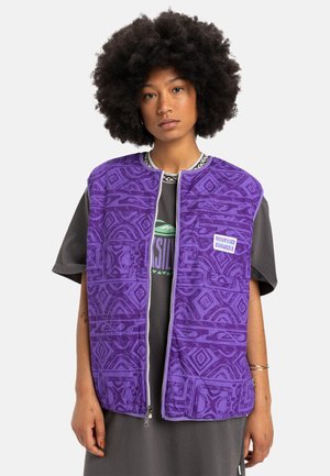 Purple padded vest featuring intricate geometric patterns, a front zipper, and two pockets. Worn over a gray t-shirt with a graphic design.