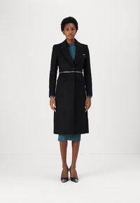 Cappotto lungo nero con chiusura a un solo bottone e dettaglio in cinturino argento. Indossato sopra un abito a coste verde acqua, accompagnato da décolleté nere a punta.
