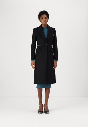 Cappotto lungo nero con chiusura a un solo bottone e dettaglio in cinturino argento. Indossato sopra un abito a coste verde acqua, accompagnato da décolleté nere a punta.
