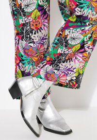 Kleurrijke, bedrukt broek met tropische motieven en tekst, gecombineerd met glanzende zilveren cowboylaarzen met een blokhak en een spitse neus.